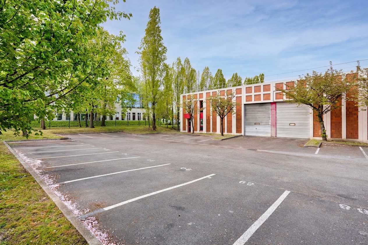 Un parking vide avec des places délimitées, situé devant un immeuble moderne aux grandes baies vitrées, entouré d'arbres et de verdure sous un ciel dégagé.