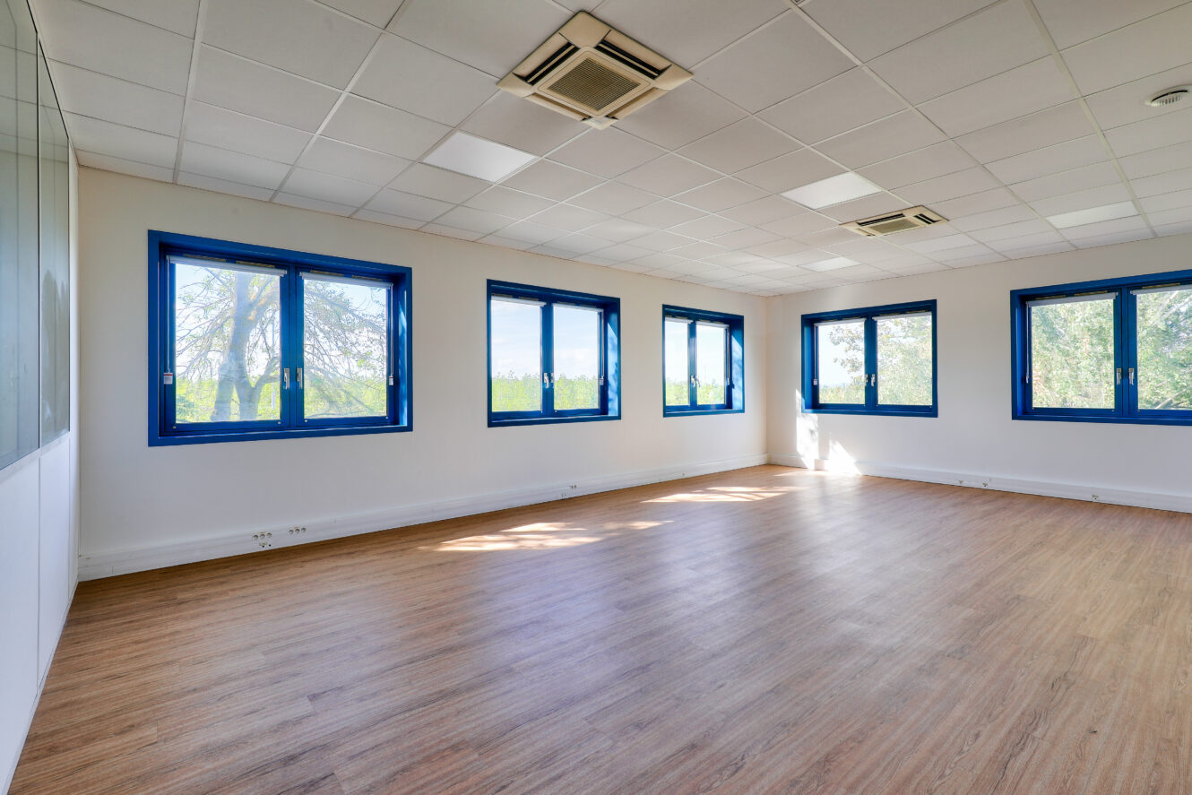 Une pièce vide avec un sol en bois, des murs blancs, des fenêtres à cadre bleu et un faux plafond blanc avec des climatiseurs montés au plafond.