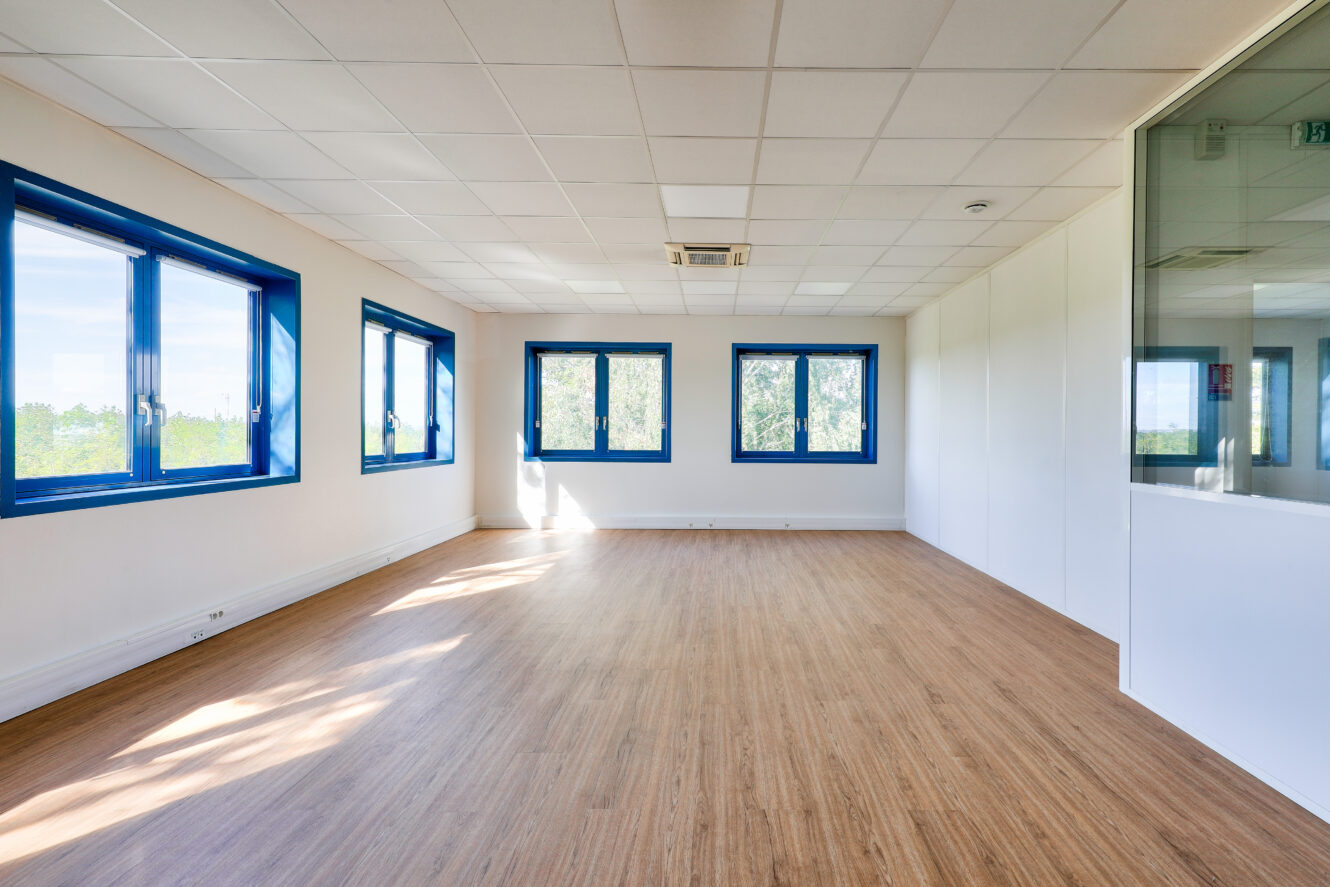 Bureau vide avec un sol en bois, des murs blancs, des fenêtres à cadre bleu et une cloison en verre sur le côté droit. La lumière naturelle pénètre par plusieurs fenêtres.