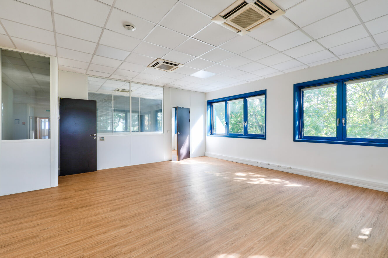 Bureau vide avec sol en bois, murs blancs, fenêtres bordées de bleu, cloison en verre et plafonniers, baigné par la lumière naturelle des grandes fenêtres.