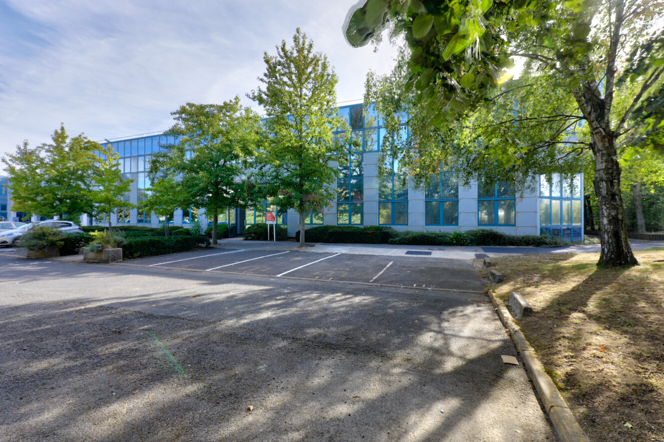 Parking vide devant un immeuble de bureaux moderne avec des baies vitrées, entouré d'arbres verts par une journée ensoleillée.