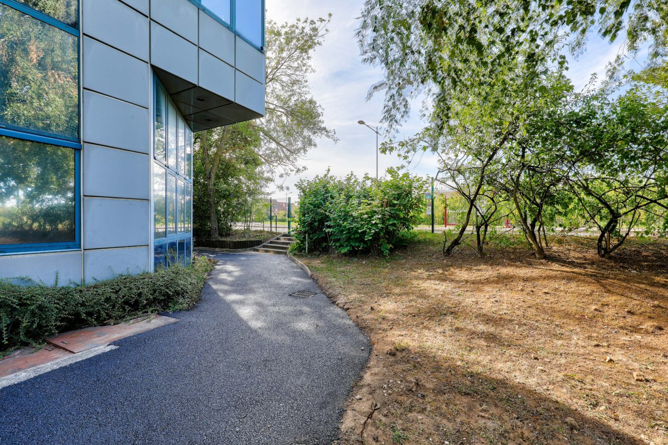 Un chemin pavé longe un bâtiment moderne en verre, bordé d'arbustes et d'arbres, avec une pelouse et des marches visibles à l'arrière-plan.