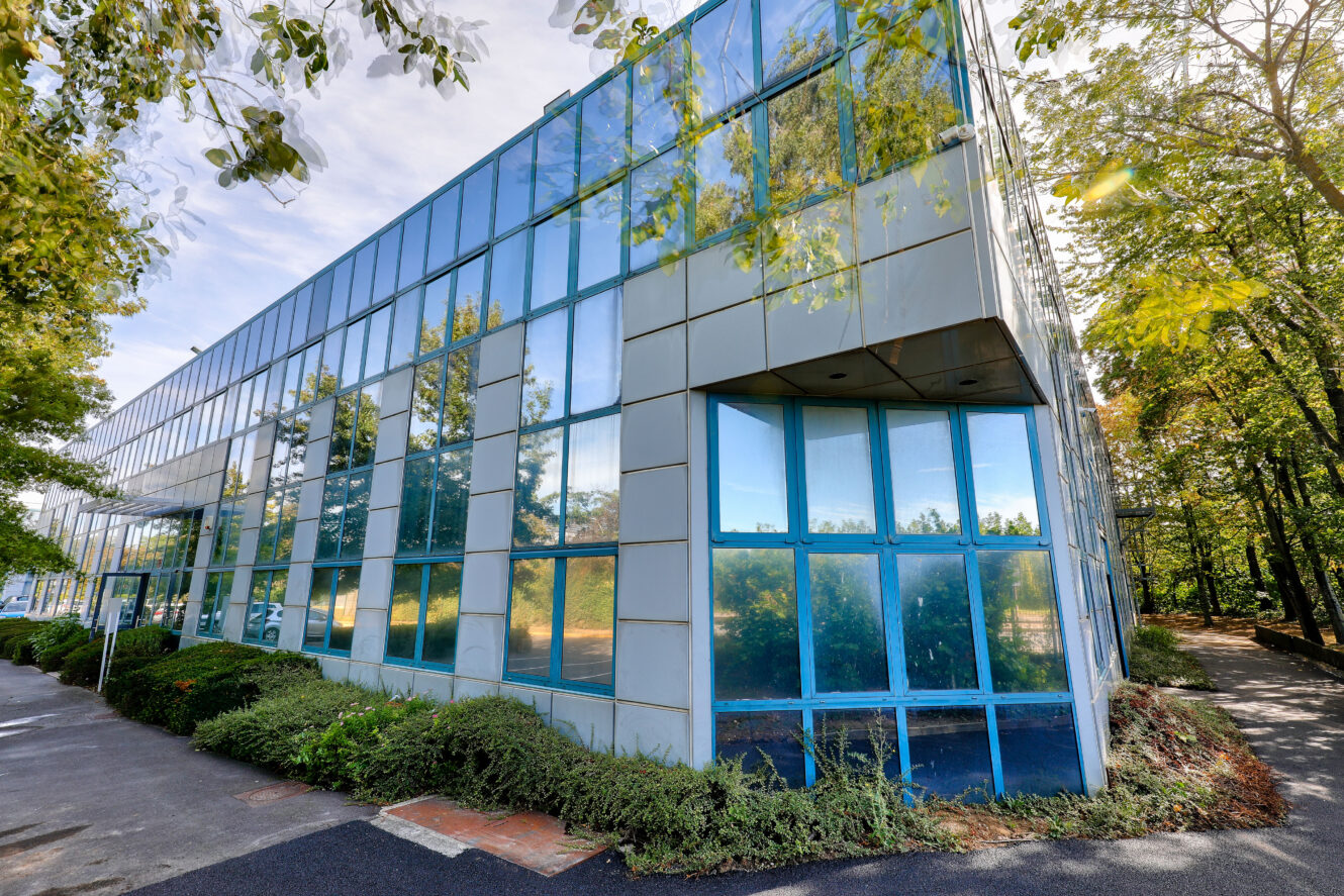 Un immeuble de bureaux moderne en verre avec des fenêtres teintées de bleu, entouré de verdure et d'arbres sous un ciel partiellement nuageux.