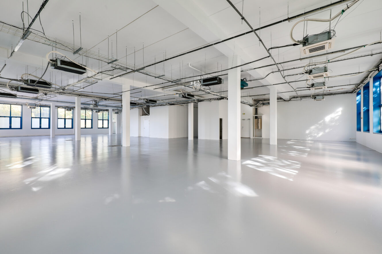 Pièce spacieuse et vide de style industriel avec des sols blancs, des tuyaux de plafond apparents, de grandes fenêtres et beaucoup de lumière naturelle.