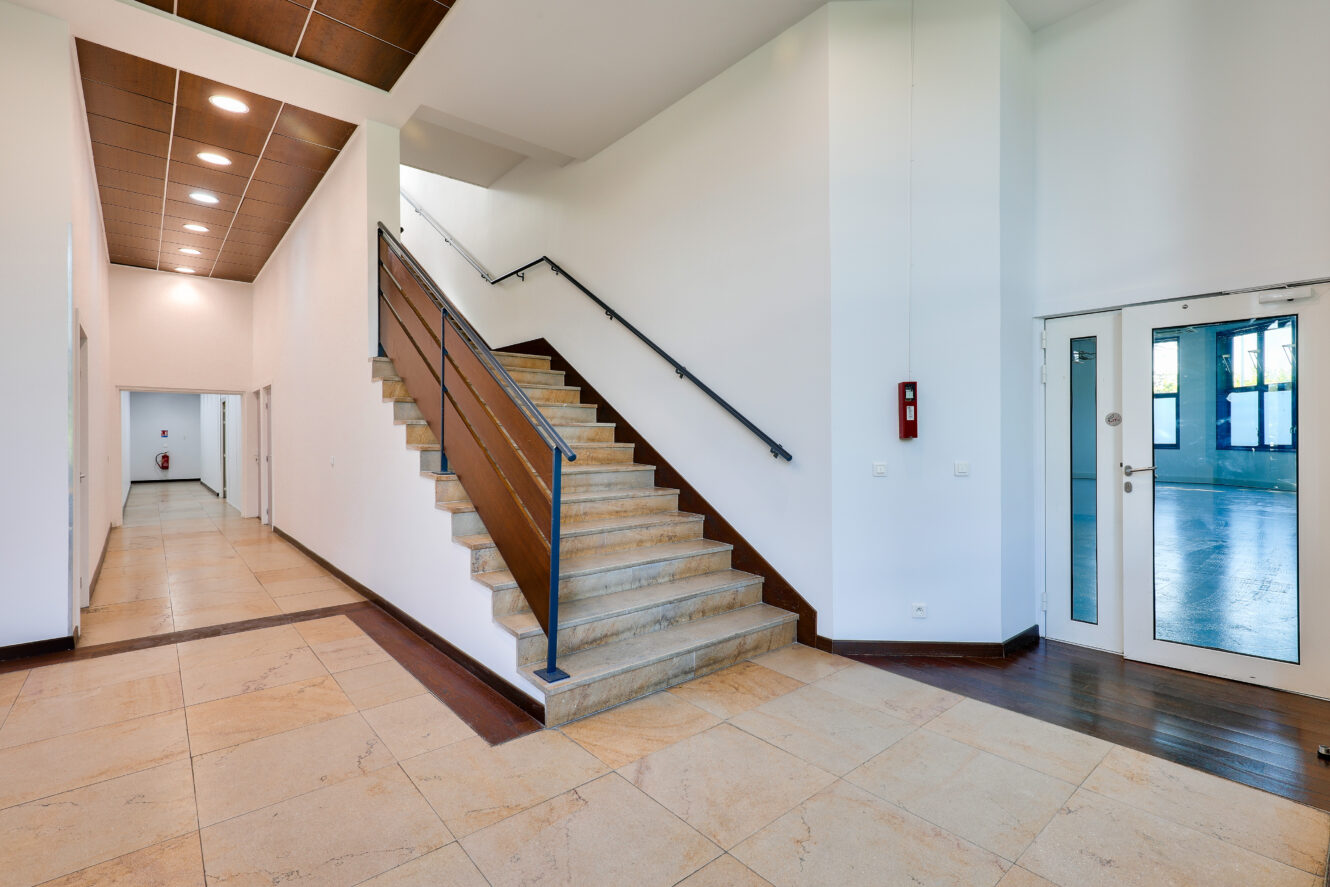 Intérieur d'un bâtiment moderne avec des sols carrelés, un escalier central avec des rampes en métal, des murs blancs, un couloir à gauche et des doubles portes en verre à droite.