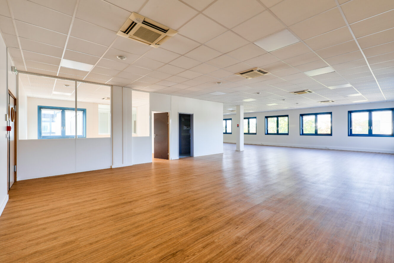 Bureau spacieux et vide avec un sol en bois, des murs blancs, des dalles de plafond, plusieurs fenêtres et une pièce vitrée dans le coin.