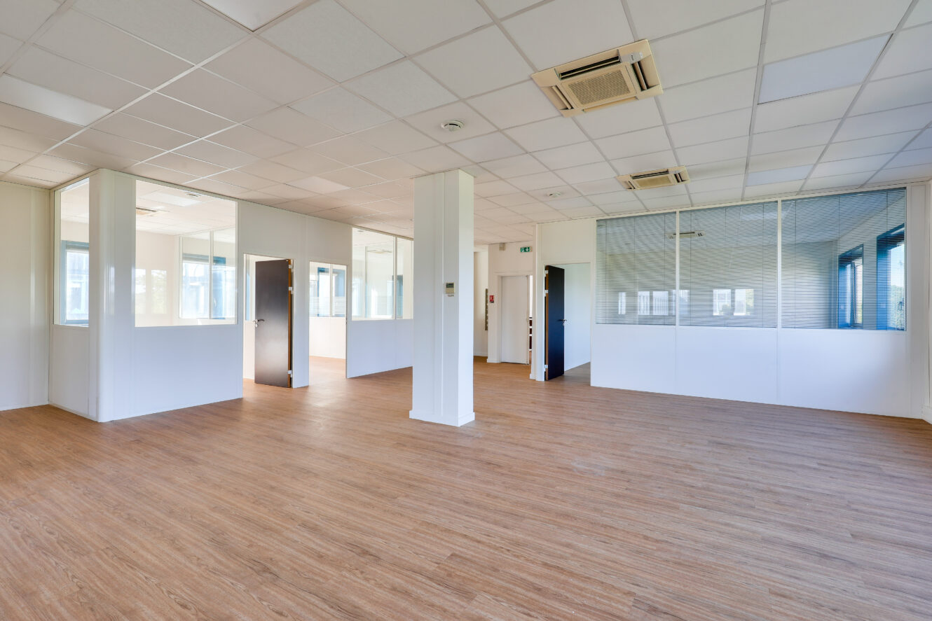 Bureau spacieux et vide avec un sol en bois, des murs blancs, des cloisons en verre, plusieurs portes et un plafond avec des lampes fluorescentes et des climatiseurs.