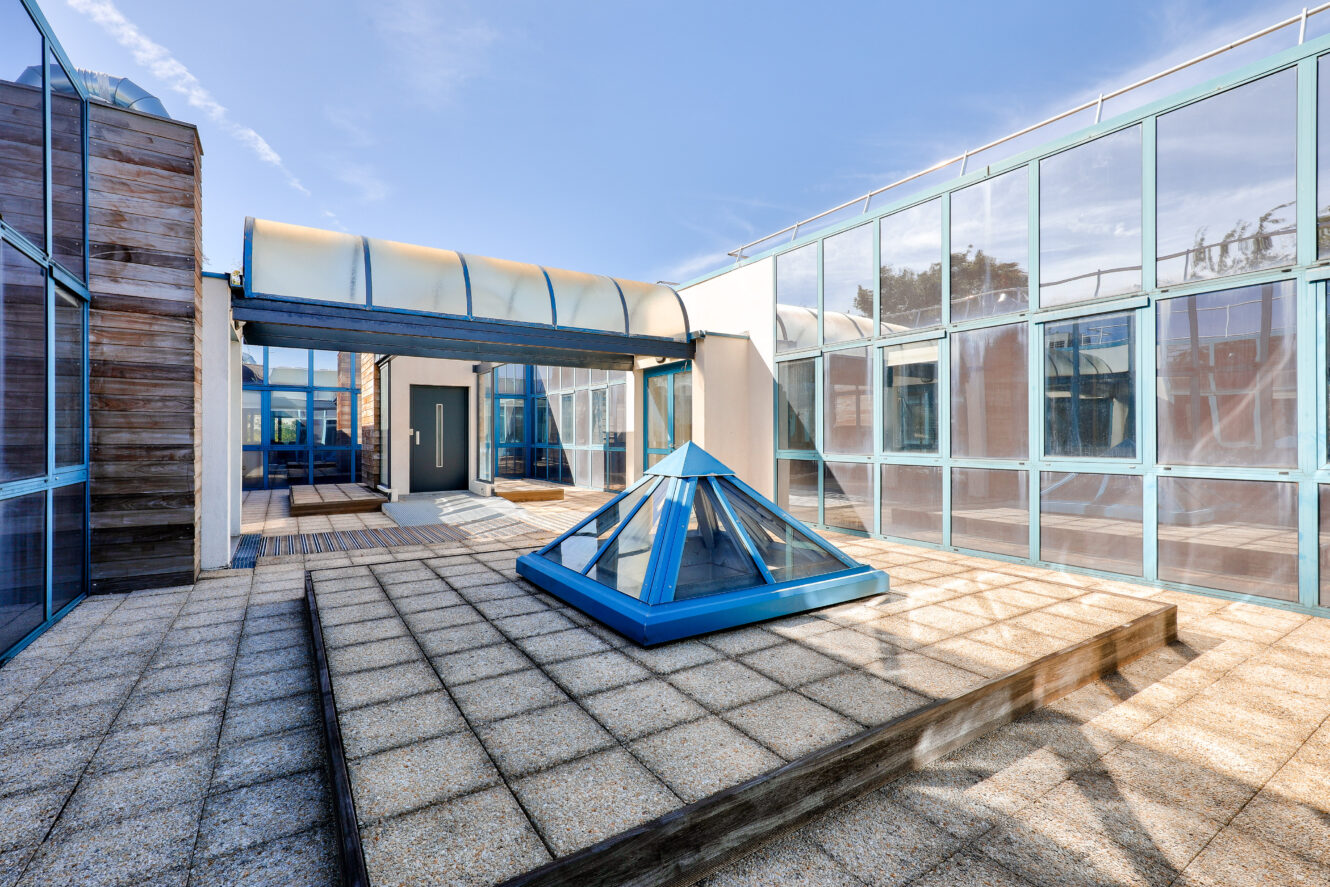 Un toit-terrasse moderne avec des fenêtres en verre, un puits de lumière pyramidal en verre bleu au centre et un pont piétonnier reliant deux sections du bâtiment sous un ciel dégagé.