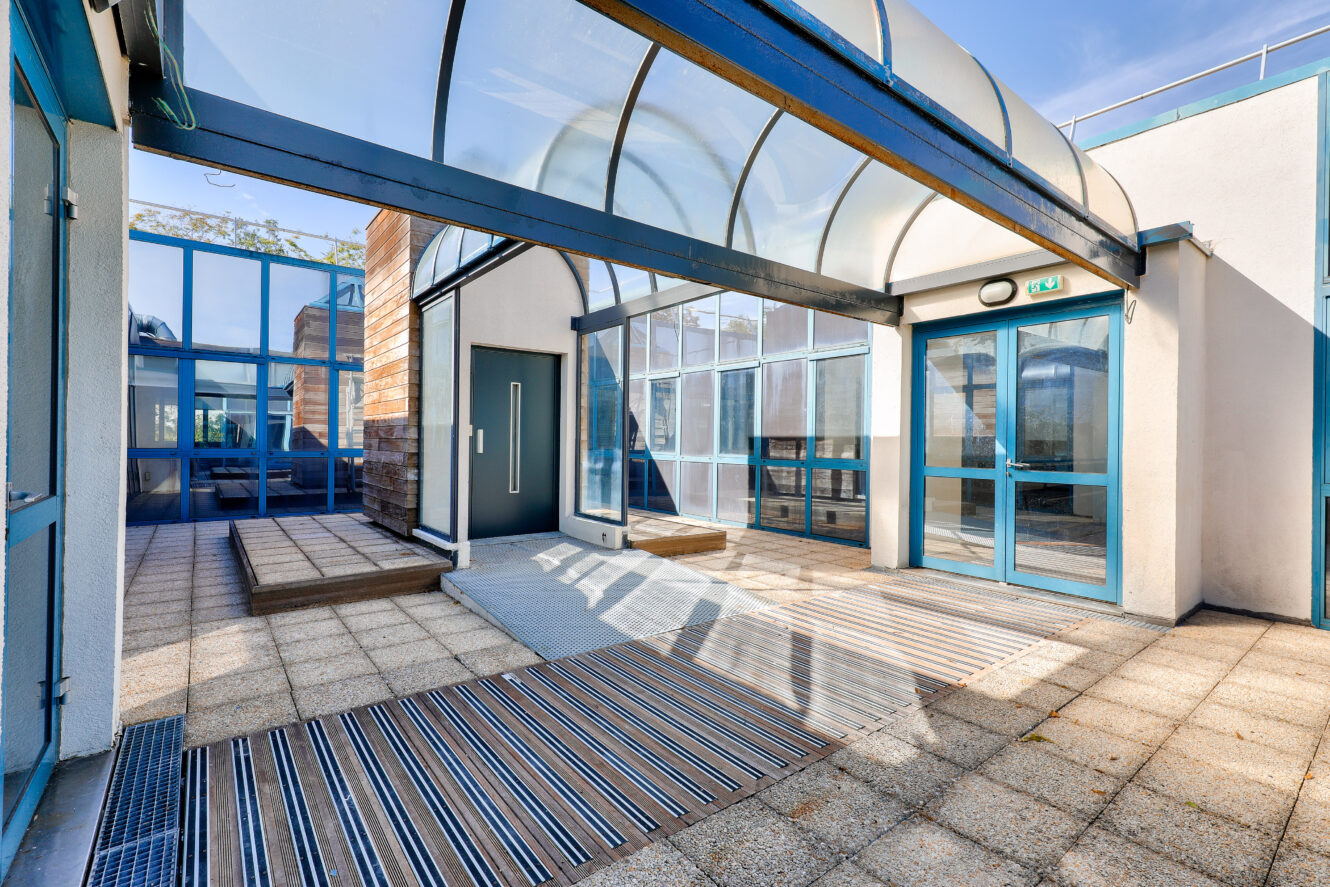 Cour d'un bâtiment moderne avec des portes en verre, des cadres métalliques bleus et un auvent arqué transparent au-dessus d'une journée ensoleillée.