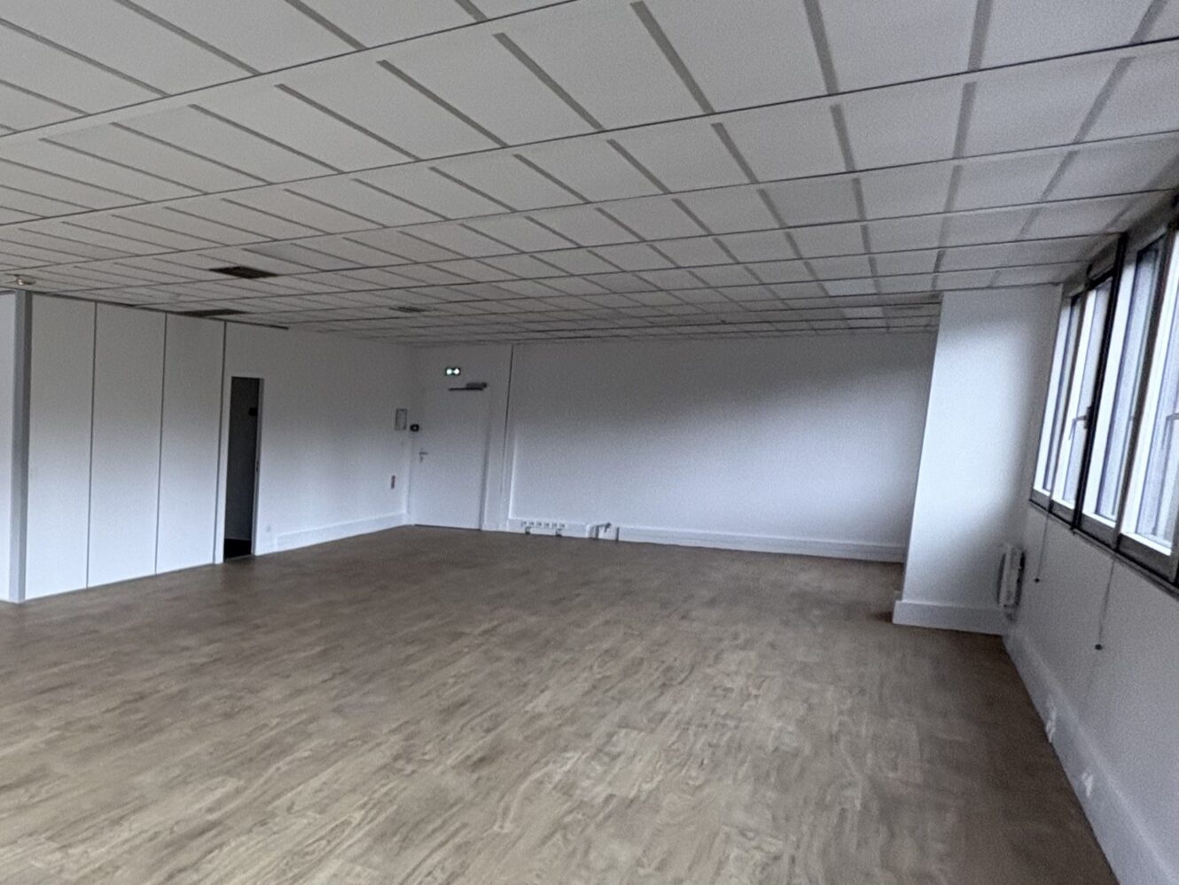 Pièce vide avec un sol en bois clair, des murs blancs, un faux plafond et des fenêtres sur le côté droit. Une porte et des panneaux utilitaires sont visibles sur le mur de gauche.