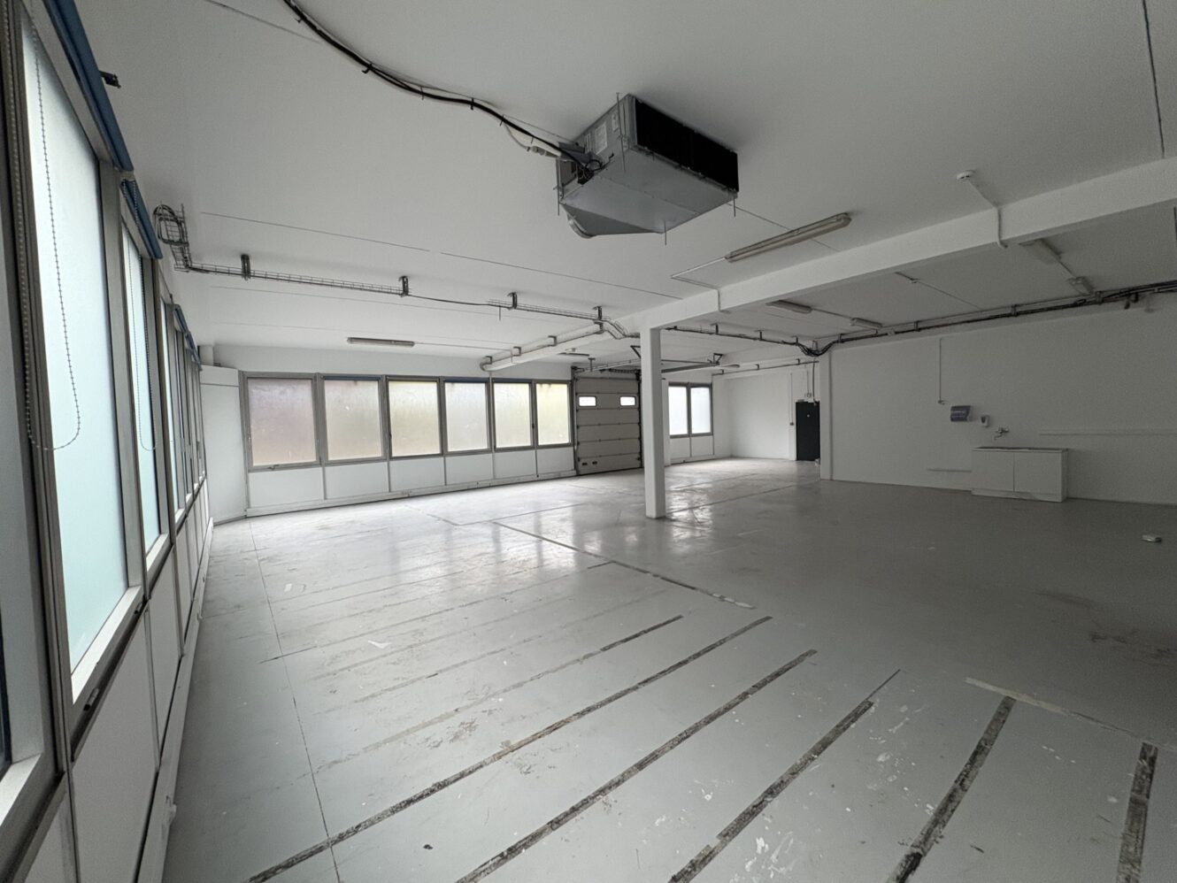 Garage industriel vide avec des murs blancs, de grandes fenêtres sur la gauche, des plafonniers, des câbles visibles, une bouche d'aération au plafond, un évier et une porte de garage fermée.