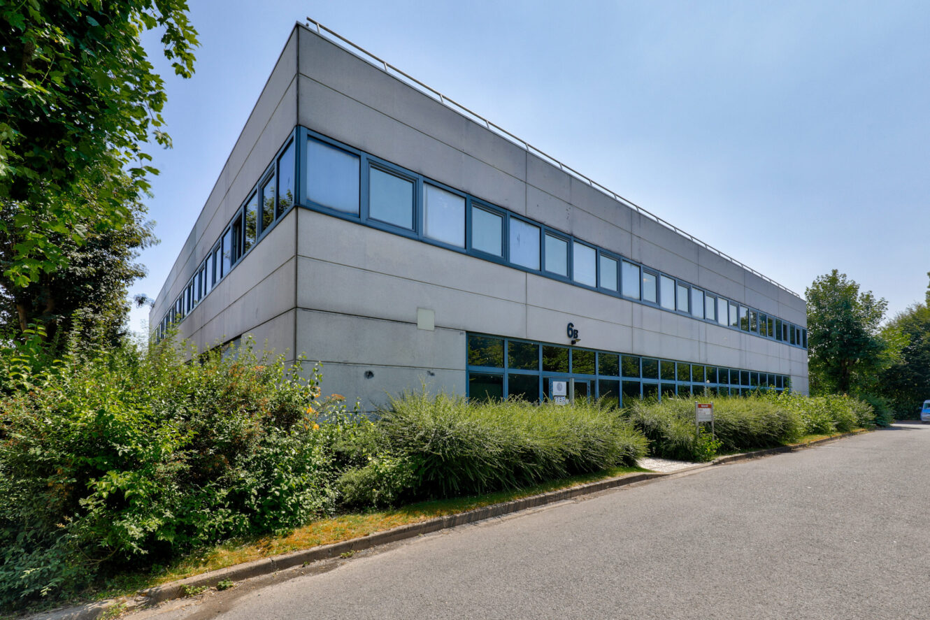 Un immeuble de bureaux moderne de deux étages avec de grandes fenêtres, entouré de verdure et de buissons, par temps clair.