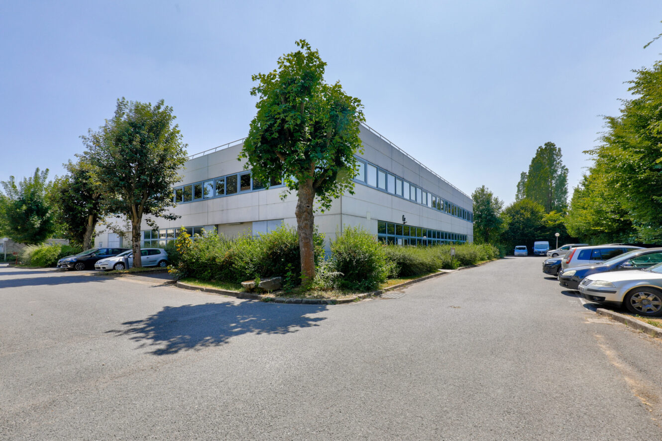 Un immeuble de bureaux moderne de deux étages avec de grandes fenêtres, entouré de verdure et d'un parking avec plusieurs voitures garées lors d'une journée ensoleillée.