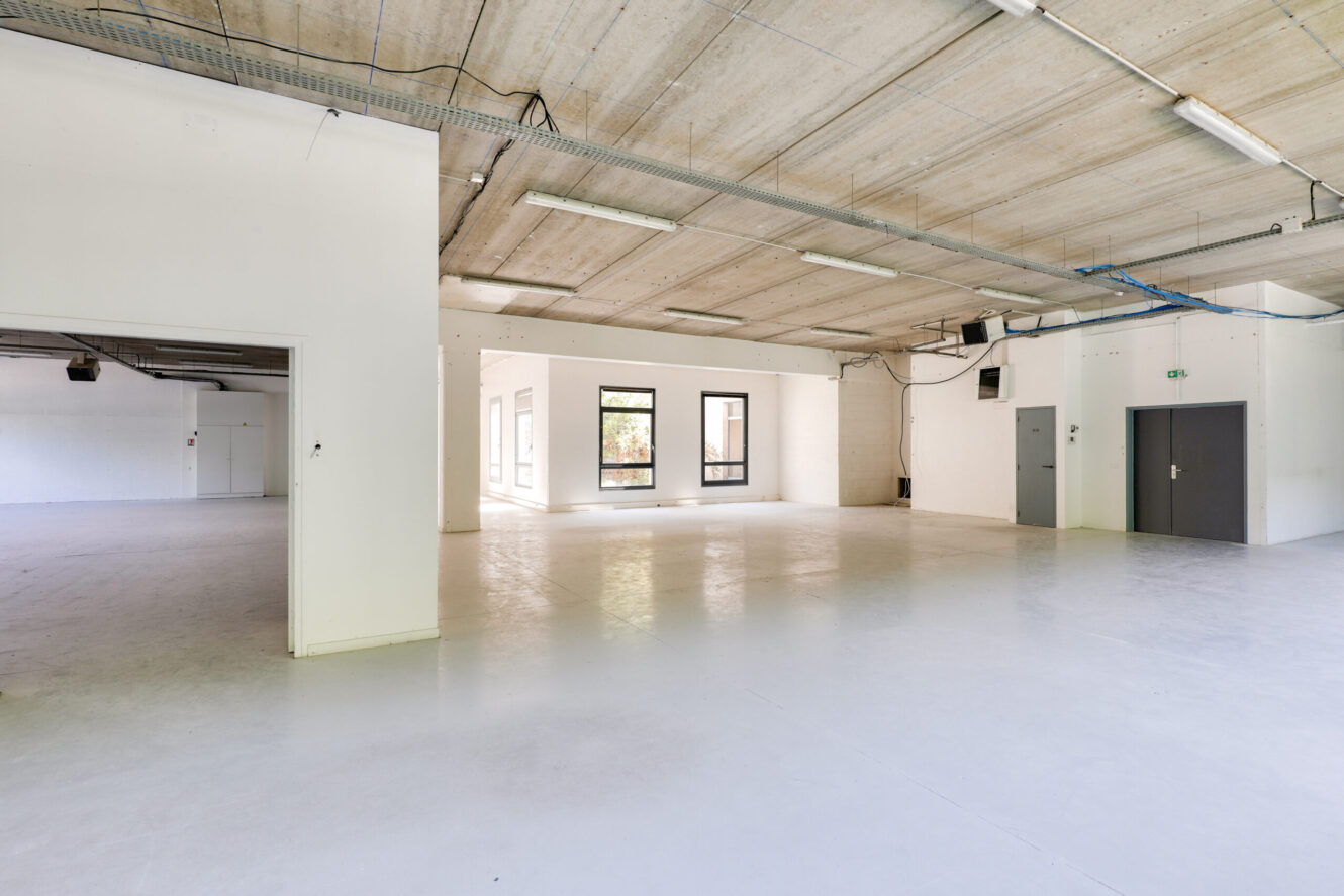 Pièce industrielle spacieuse et vide avec des murs blancs, de grandes fenêtres, un plafond en béton, des câbles apparents et un éclairage fluorescent.