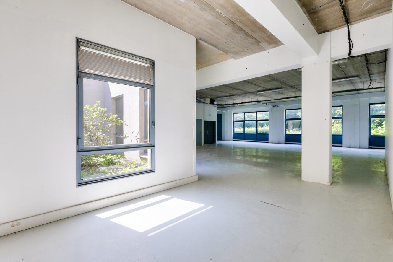 Pièce spacieuse et vide de style industriel avec des murs blancs, de grandes fenêtres, un plafond en béton et une lumière naturelle qui pénètre à l'intérieur.