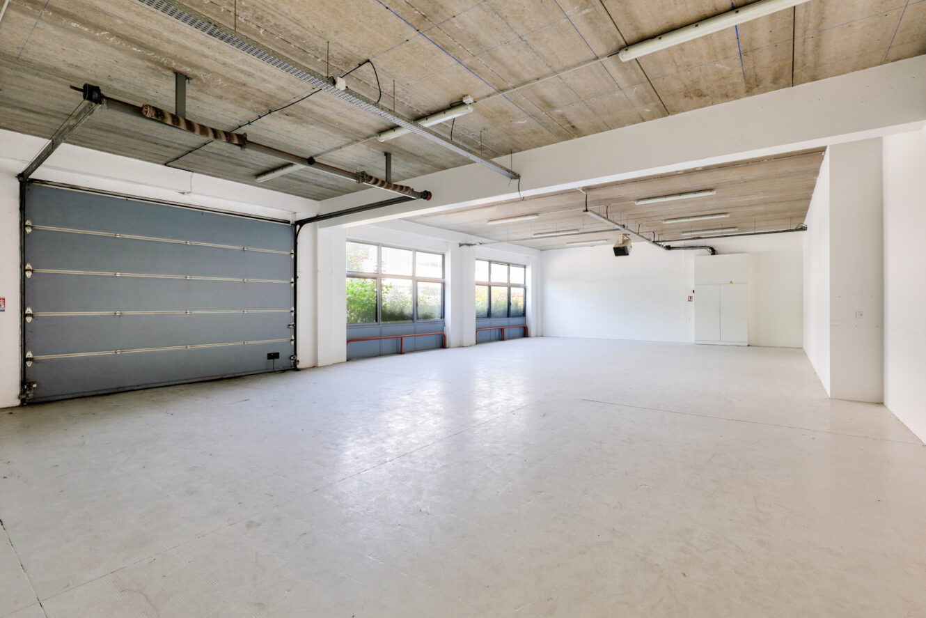Garage vide et spacieux avec un sol et un plafond en béton, de grandes fenêtres, des murs blancs et une porte roulante fermée. Des lampes fluorescentes sont montées au plafond.