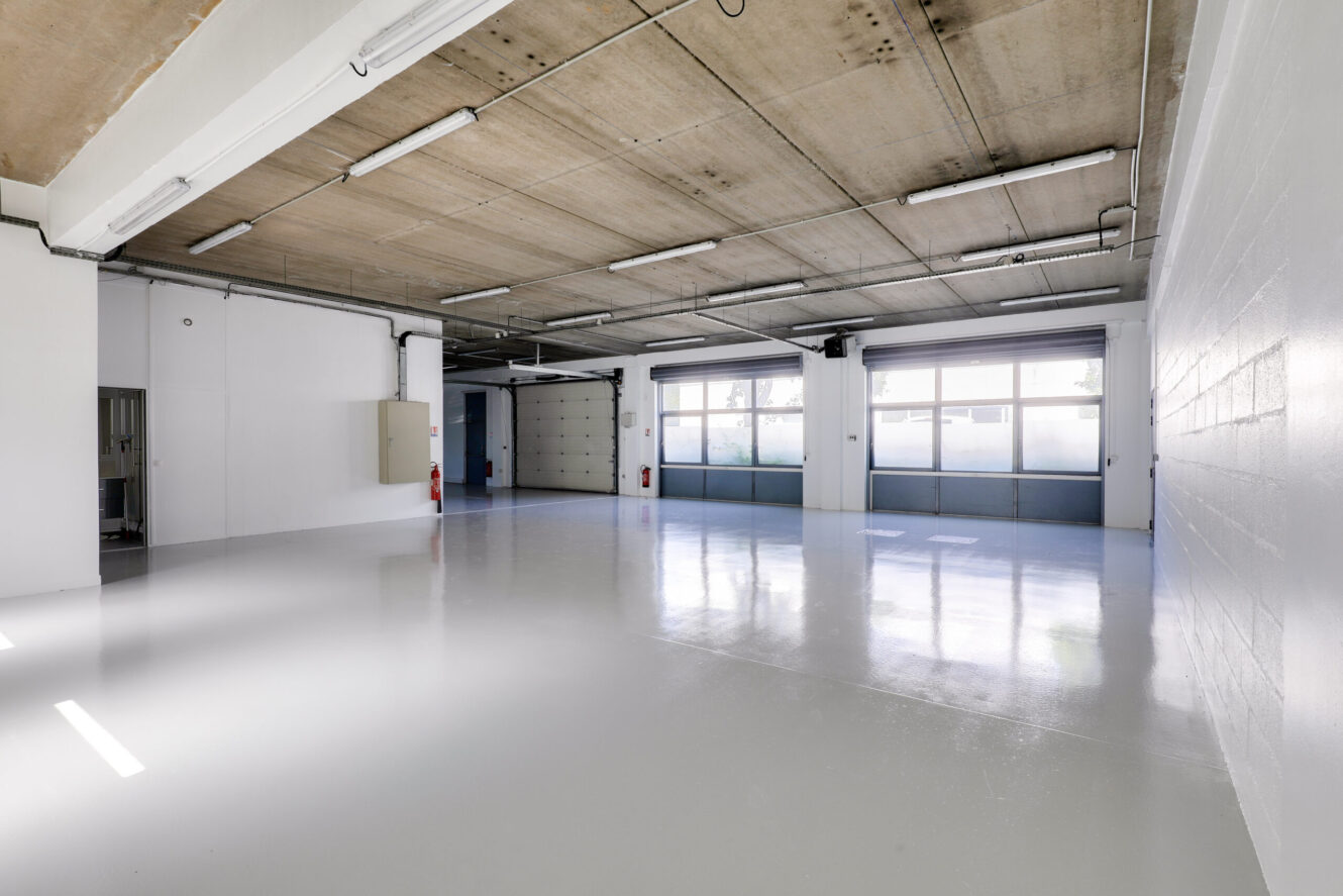 Pièce industrielle spacieuse et vide, avec des sols polis, des murs blancs, de grandes fenêtres et un plafond en béton apparent, éclairée par des lampes fluorescentes.