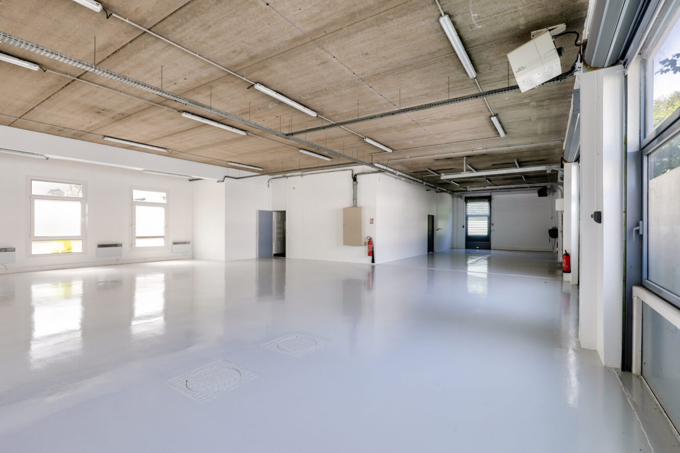 Pièce industrielle spacieuse et vide avec un sol blanc poli, un plafond en béton, des lumières fluorescentes et de grandes fenêtres le long d'un mur.