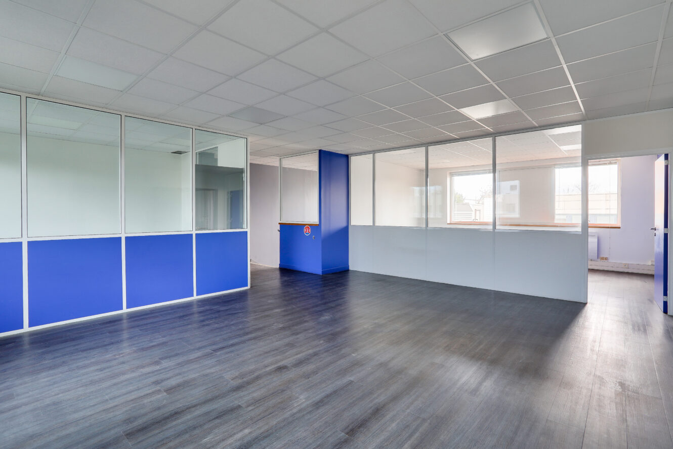 Bureau moderne vide avec des cloisons en verre, des murs bleus et blancs, une grande fenêtre et un sol en bois foncé.