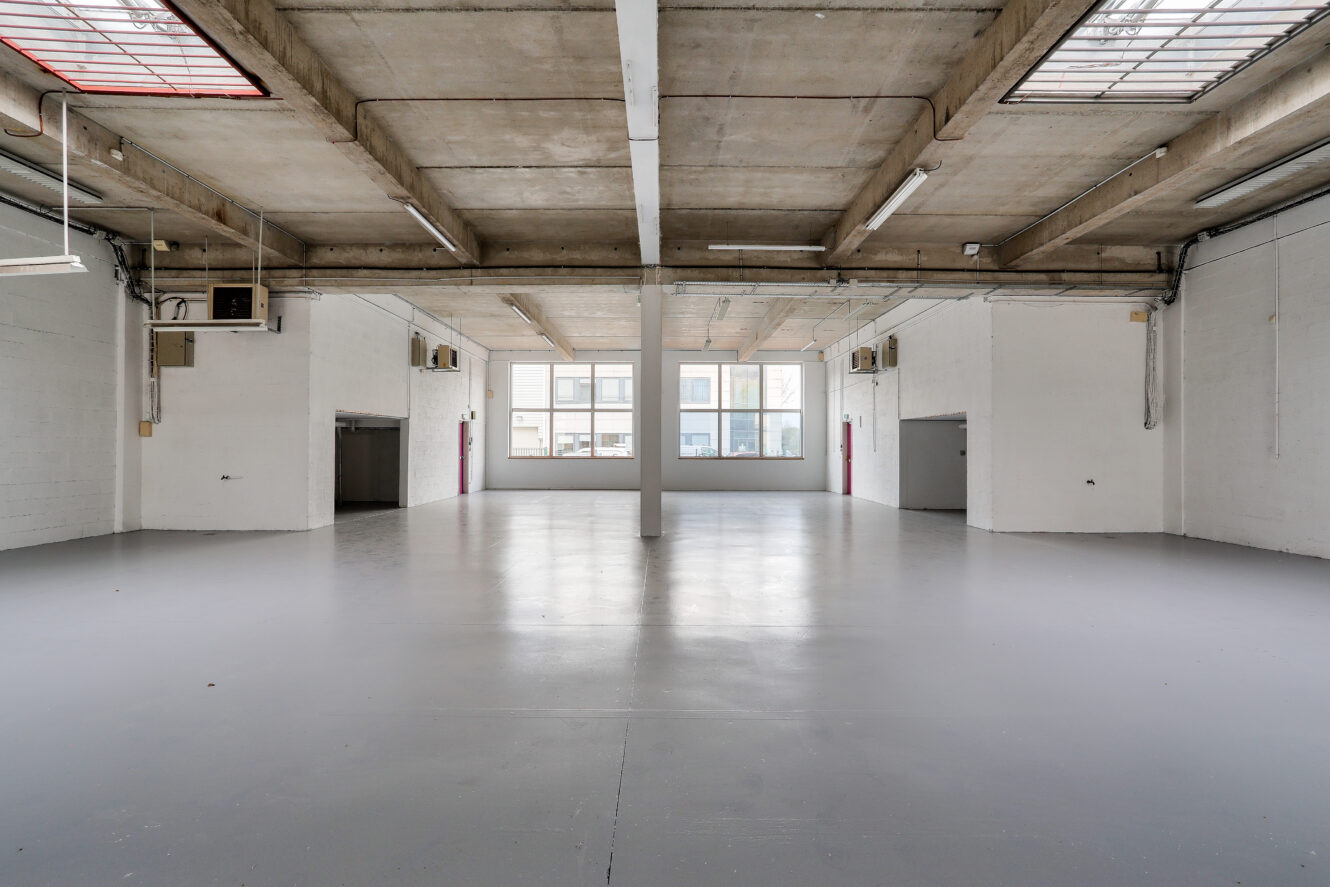 Un entrepôt industriel vide avec des sols en béton, des poutres apparentes au plafond, de grandes fenêtres et des murs blancs.