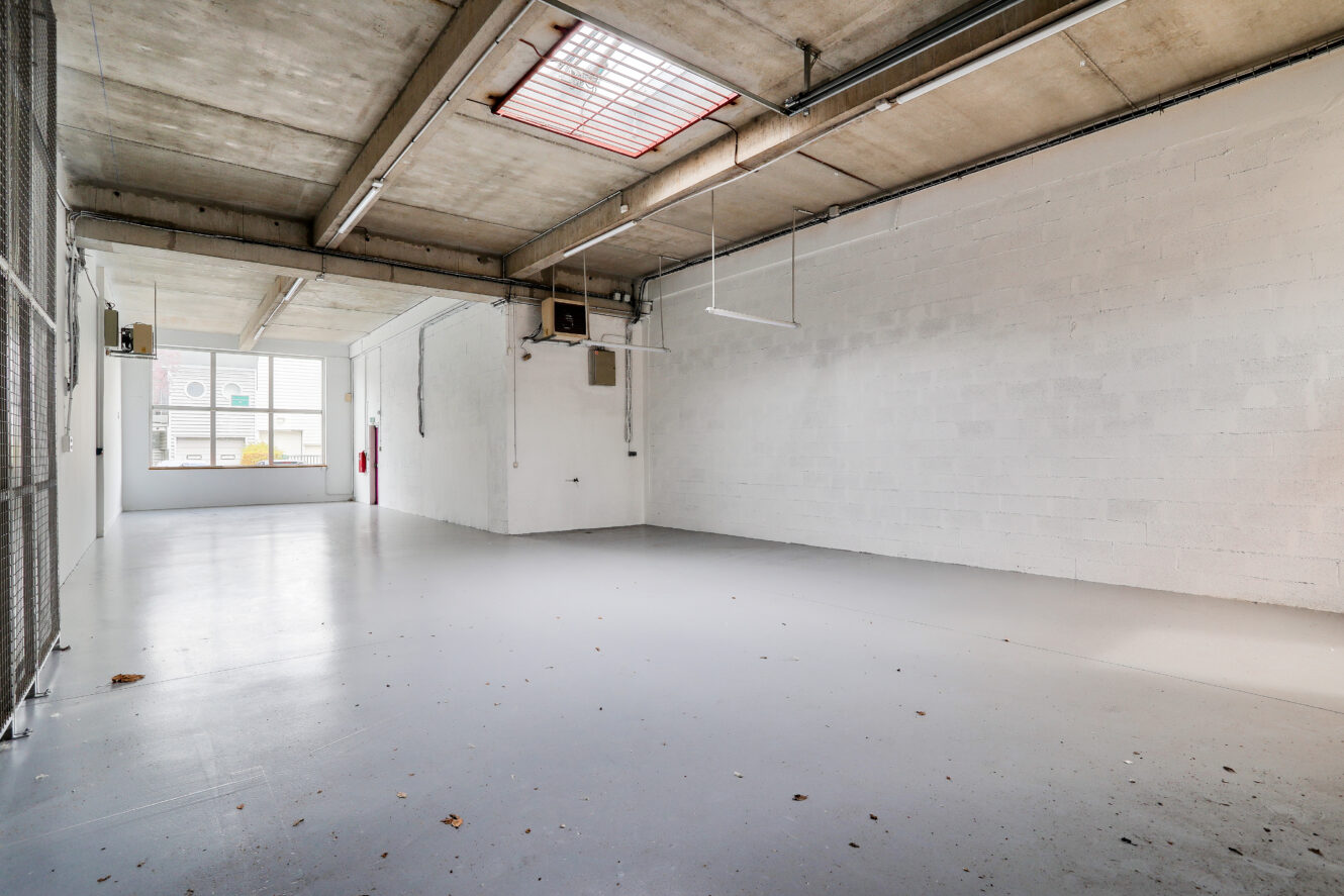 Un entrepôt industriel vide avec des sols en béton, des plafonds apparents, des murs en briques peintes en blanc, des lampes fluorescentes et une grande fenêtre à l'extrémité qui laisse entrer la lumière naturelle.