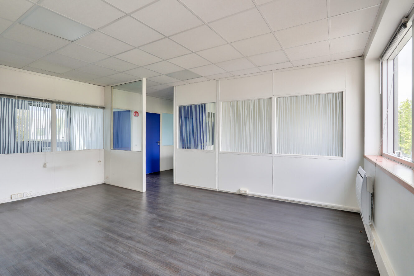 Bureau vide avec de grandes fenêtres, des murs blancs, des cloisons vitrées avec stores et un sol en bois foncé. Une porte bleue est visible à l'arrière-plan.