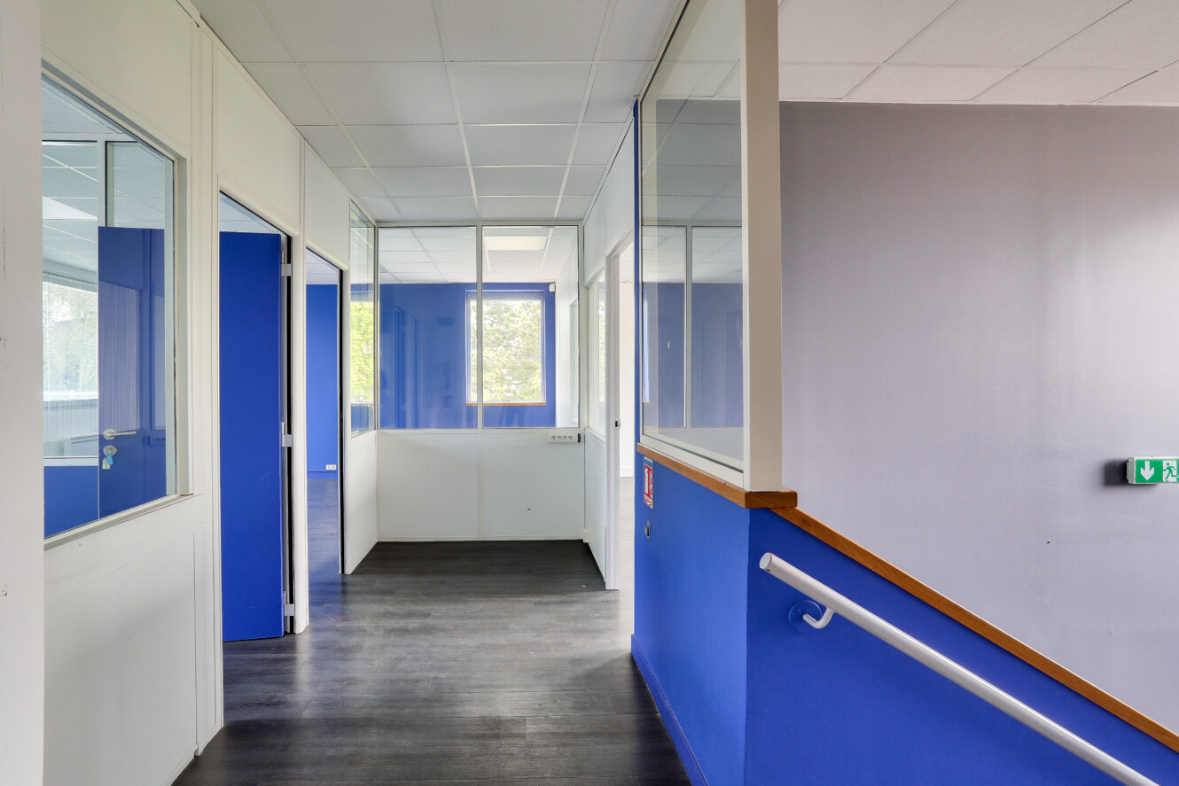 Un couloir avec des murs bleus et blancs, des cloisons de bureau en verre, un sol sombre et de la lumière naturelle provenant des fenêtres au bout du couloir.