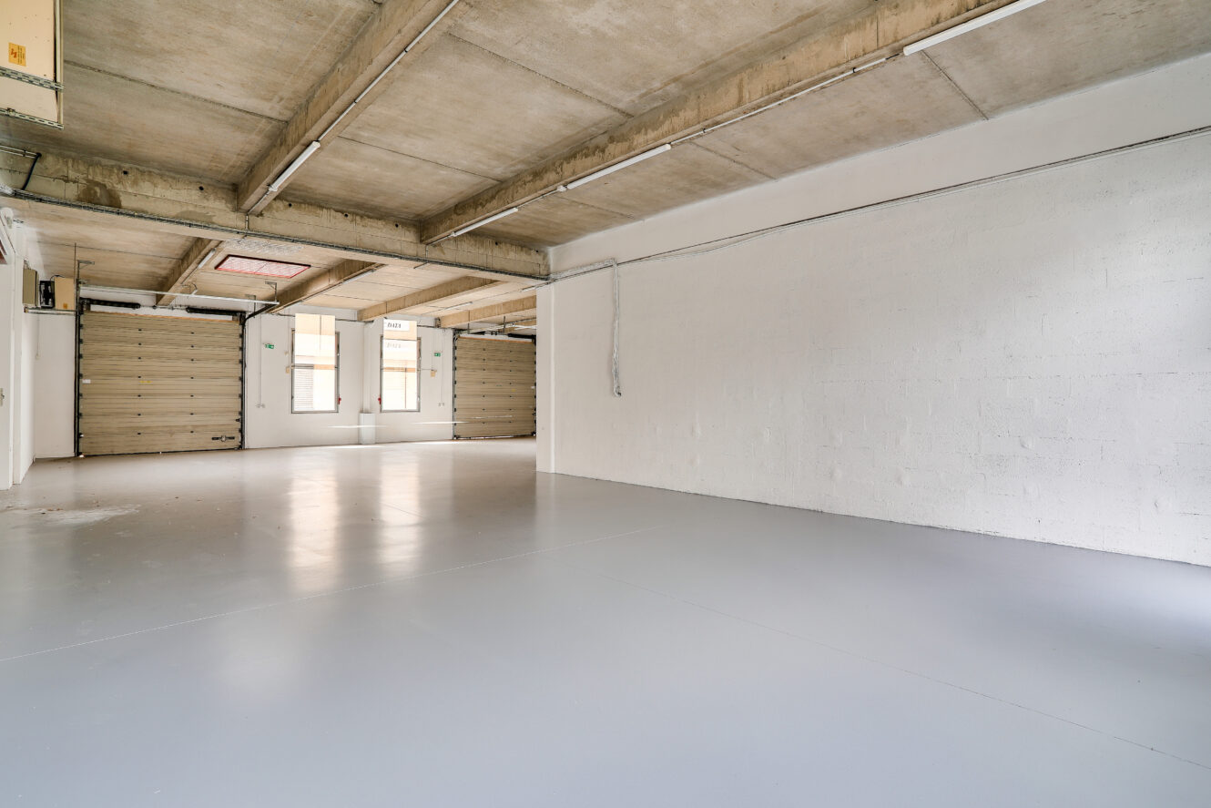 Garage industriel vide avec plafond en béton, murs peints en blanc, sol gris lisse, grandes fenêtres et deux portes roulantes fermées.