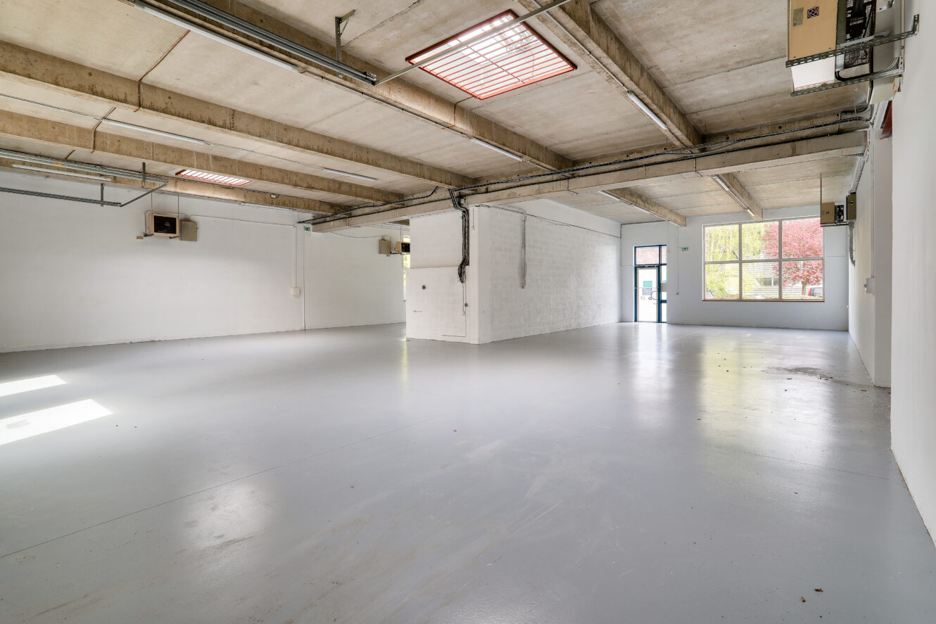 Spacieux entrepôt industriel vide avec un plafond en béton, des murs blancs et de grandes fenêtres laissant entrer la lumière naturelle. Une seule porte est visible à l'extrémité.