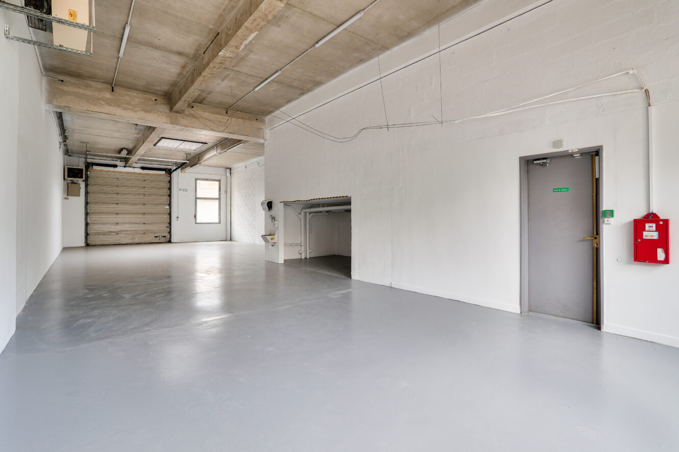 Pièce industrielle spacieuse et vide avec des murs blancs, un plafond en béton, une porte grise, un extincteur fixé au mur et une grande porte de garage à l'extrémité.
