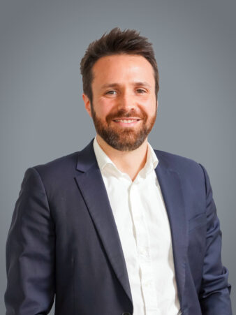 Un homme aux cheveux bruns courts et à la barbe, portant une veste de costume marine sur une chemise blanche, sourit sur un fond gris uni.