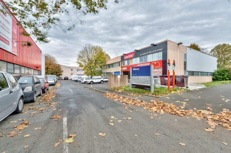 Un parking avec plusieurs voitures garées, des feuilles d'automne éparpillées sur le sol et une grande enseigne d'annuaire d'entreprises devant des immeubles de bureaux sous un ciel nuageux.