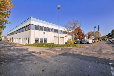 Un bâtiment commercial moderne de deux étages avec de grandes fenêtres, entouré d'arbres et de voitures garées, sous un ciel bleu clair.