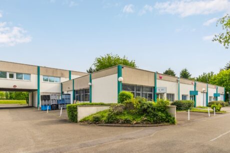 Bâtiment commercial de plain-pied avec des accents sarcelle, de grandes fenêtres et de la verdure environnante sous un ciel clair. Plusieurs caddies sont visibles près de l'entrée.
