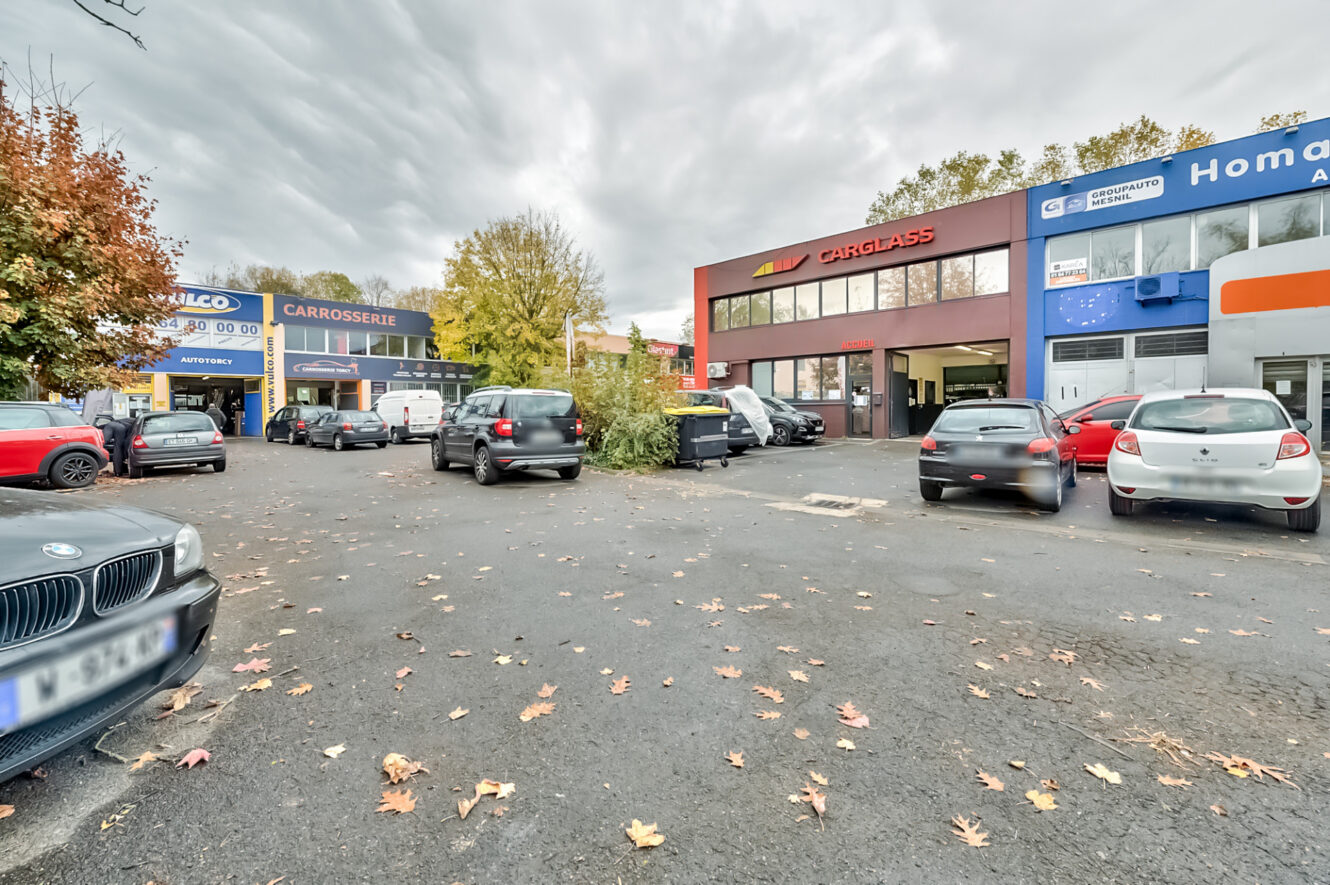 Parking avec plusieurs voitures devant des ateliers de réparation automobile et des bureaux, dont CarGlass, sous un ciel nuageux avec des arbres en arrière-plan.