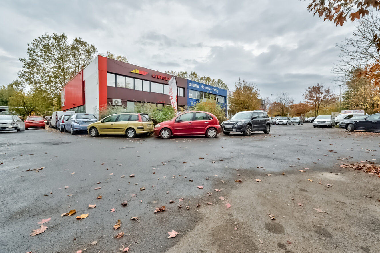 Parking avec plusieurs voitures, dont une voiture rouge au centre, devant un bâtiment de deux étages avec des panneaux rouges et blancs, par une journée nuageuse.