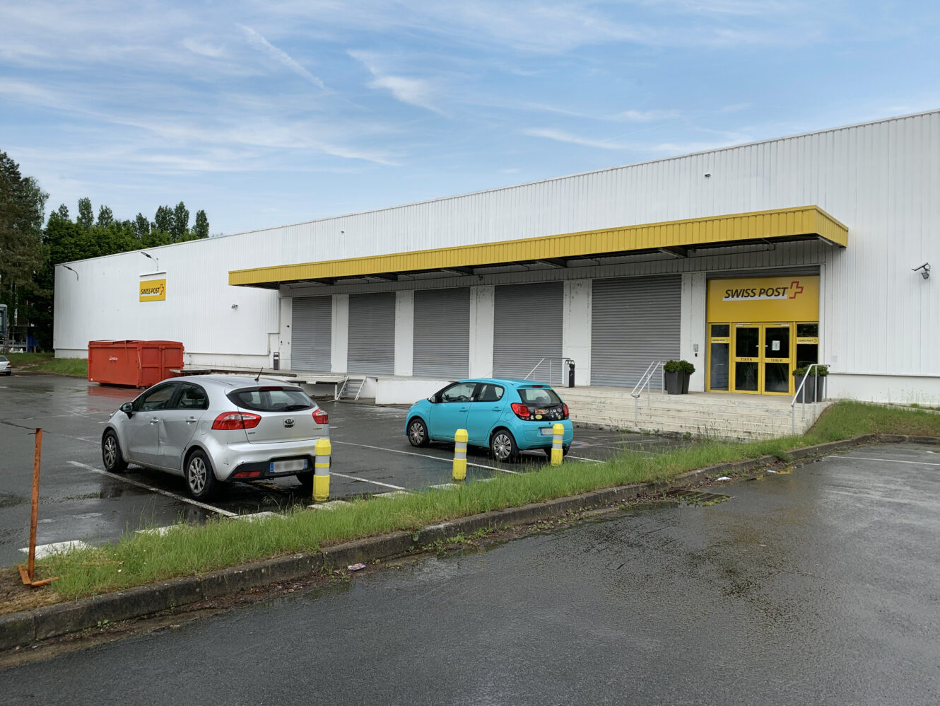 Un bâtiment de la Poste suisse avec une enseigne jaune, trois quais de chargement fermés, deux voitures garées et une chaussée mouillée au premier plan.