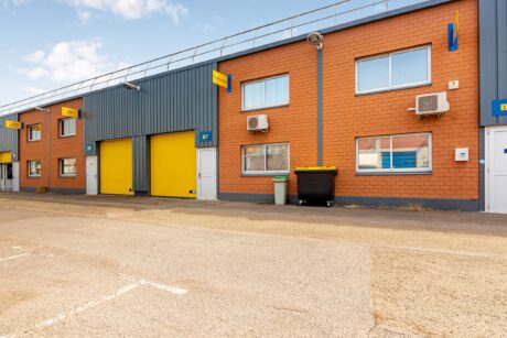 Une rangée d'unités industrielles avec des portes roulantes jaunes et des murs en briques, avec des climatiseurs et une benne noire à l'extérieur.