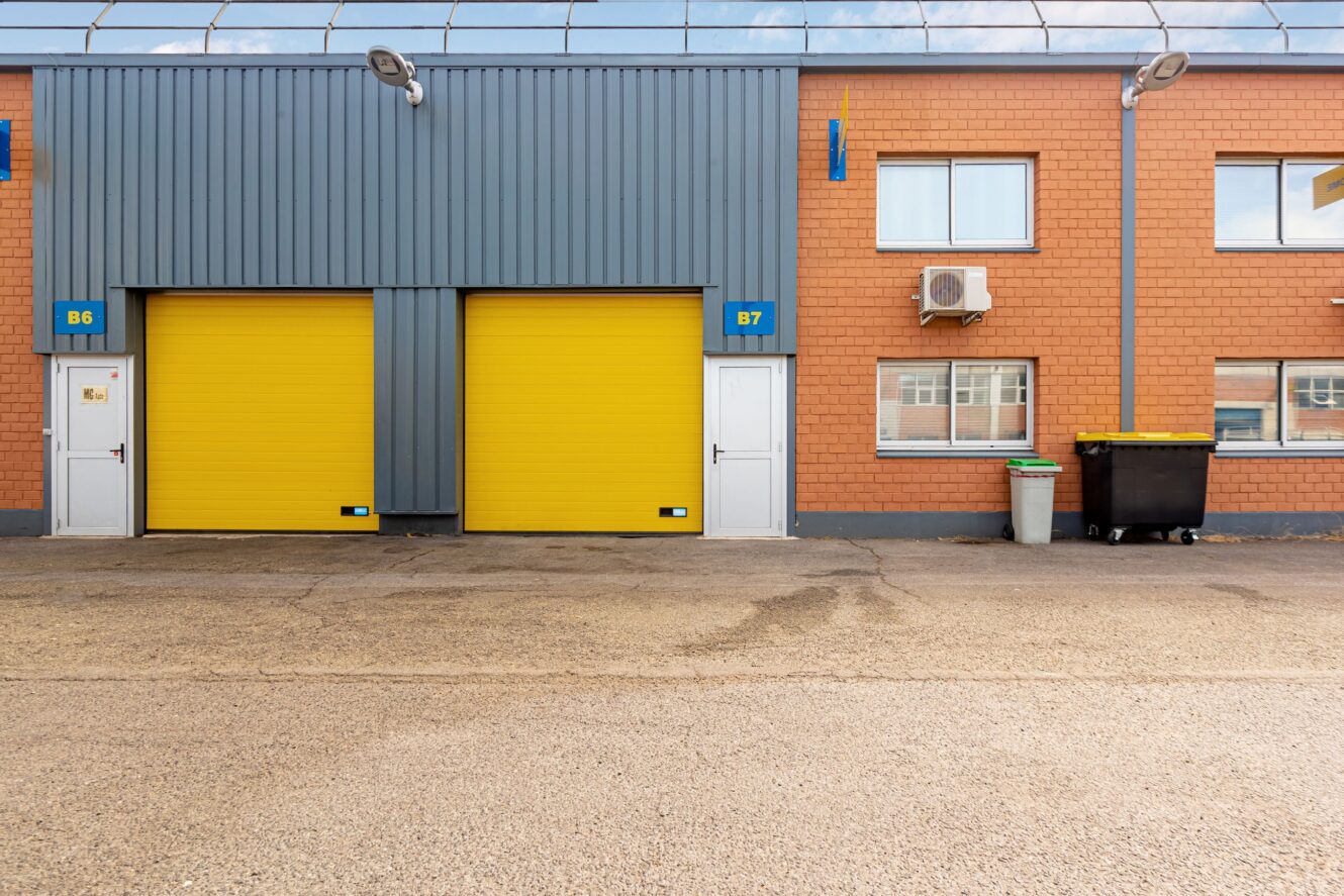 Un bâtiment commercial avec deux portes roulantes jaunes étiquetées B6 et B7, flanquées de portes blanches, de fenêtres et d'une poubelle noire à l'extérieur.