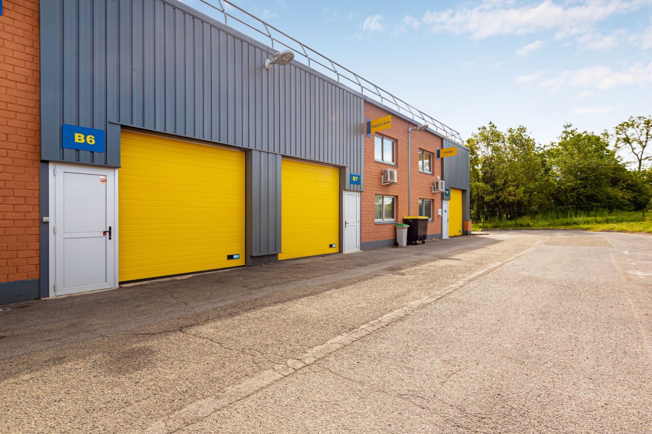 Bâtiment industriel avec deux portes roulantes jaunes et une porte blanche, marqué "B6" sur un panneau bleu, situé à côté d'un terrain pavé et d'arbres en arrière-plan.