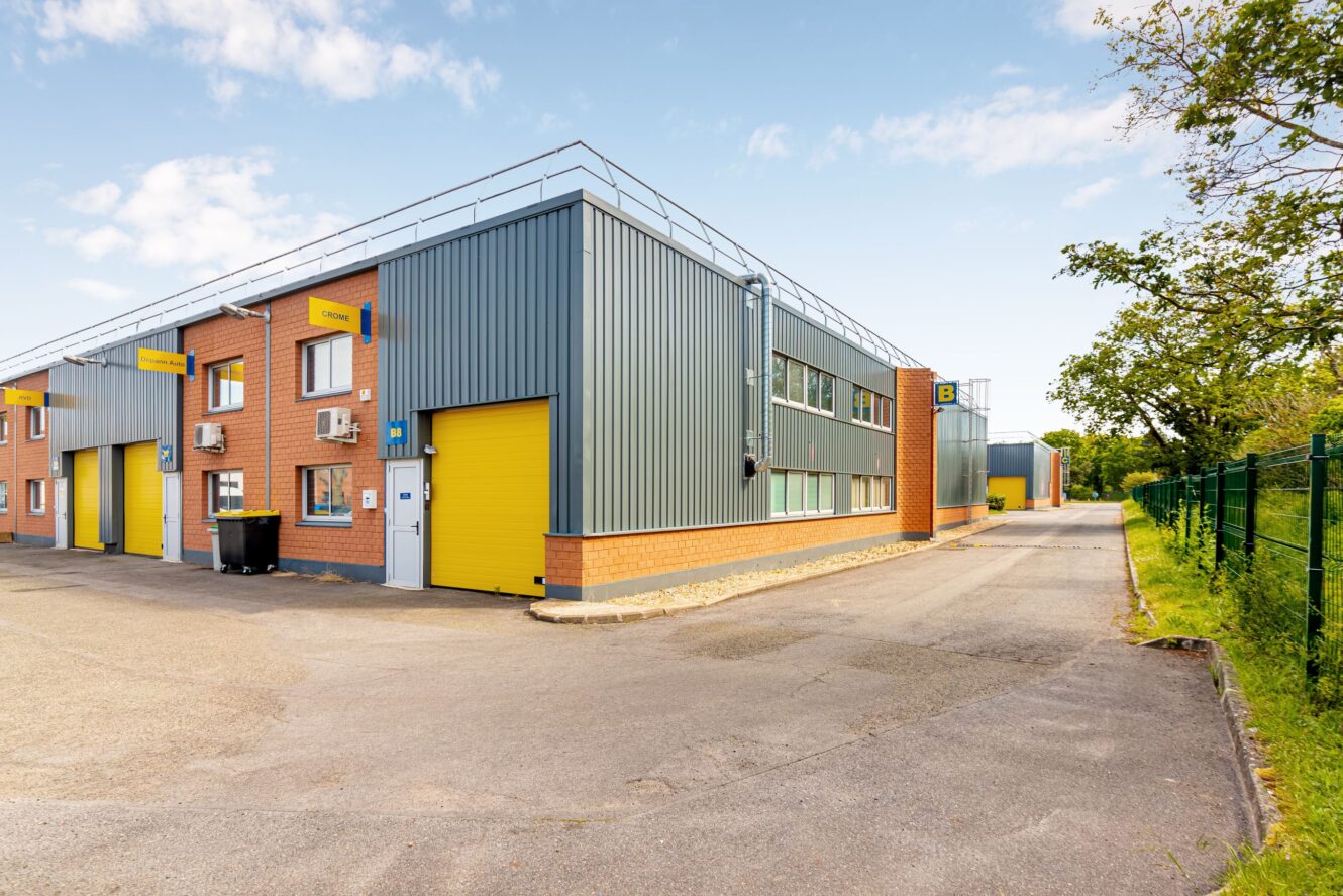 Un bâtiment industriel moderne avec un bardage métallique gris, des portes jaunes et des accents de brique, situé le long d'une route pavée avec une clôture et des arbres à proximité.