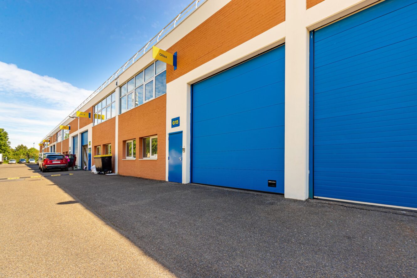 Une rangée d'unités industrielles modernes avec de grandes portes roulantes bleues et des enseignes jaunes, avec des voitures garées le long du bâtiment sous un ciel clair.