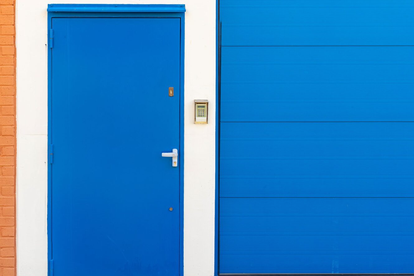 Une porte bleue avec une poignée argentée se trouve à côté d'une porte de garage bleue. Un système d'entrée à clavier est installé sur le mur blanc qui les sépare.