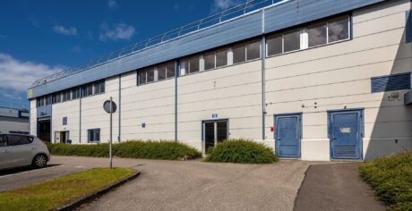 Un bâtiment industriel de deux étages avec des portes bleues, plusieurs fenêtres et un petit parking avec une voiture garée lors d'une journée ensoleillée.