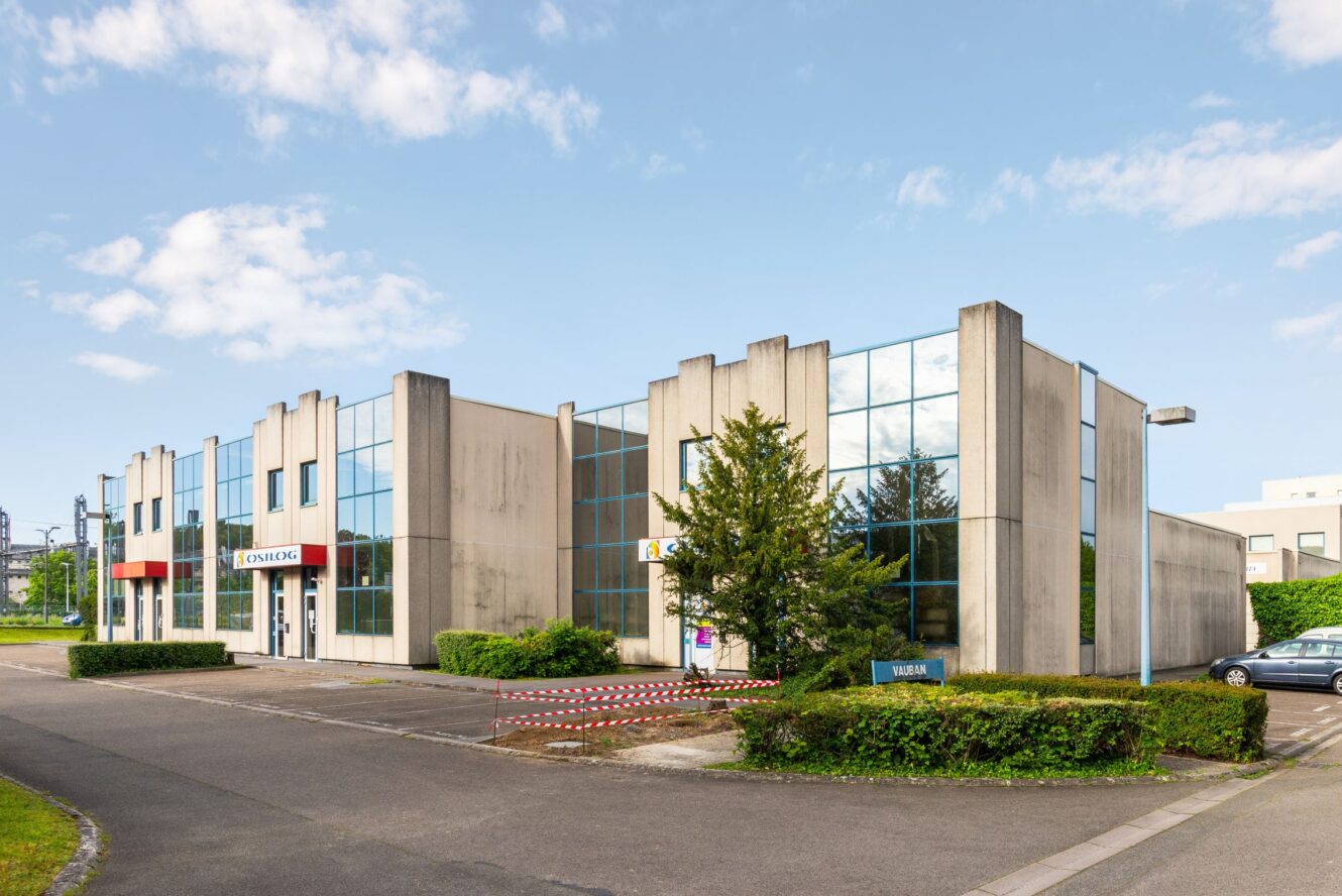 Bâtiment commercial moderne avec de grandes baies vitrées, plusieurs entrées et un petit espace paysager à l'avant, sous un ciel partiellement nuageux.