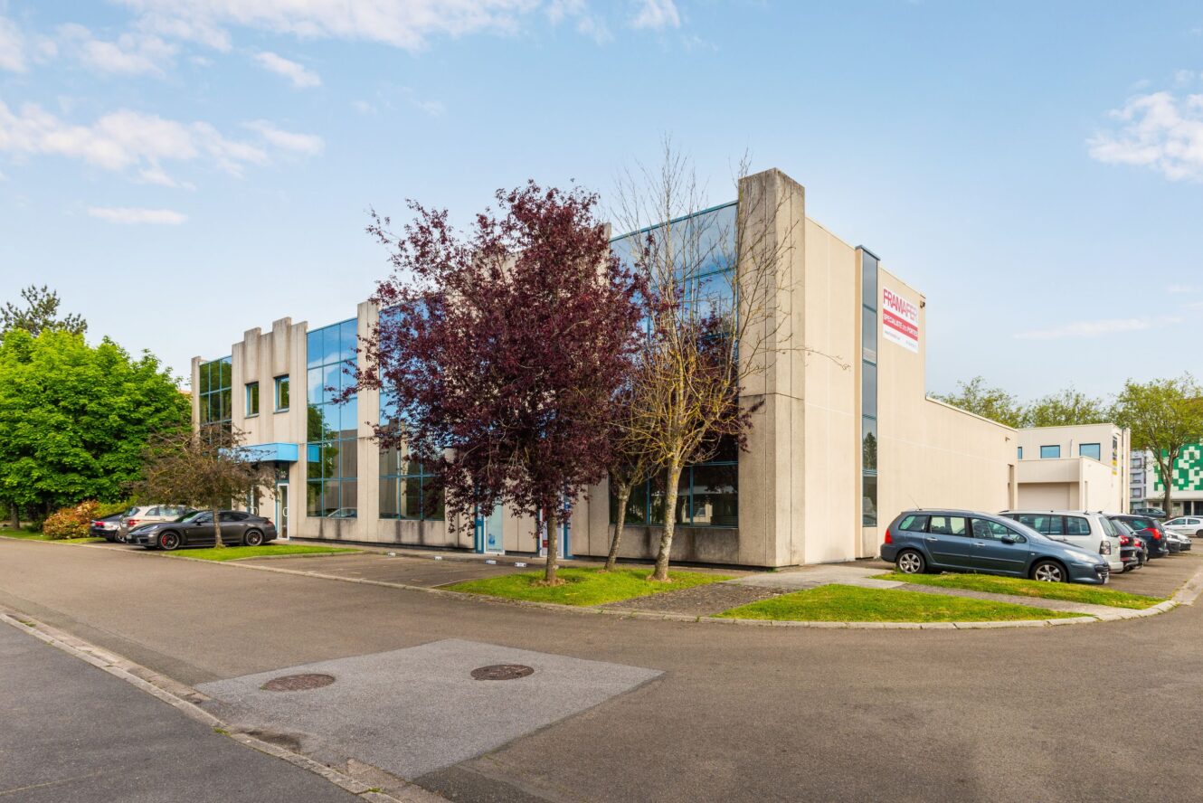 Un immeuble de bureaux de deux étages avec de grandes baies vitrées, entouré de voitures garées et d'arbres, par temps clair.