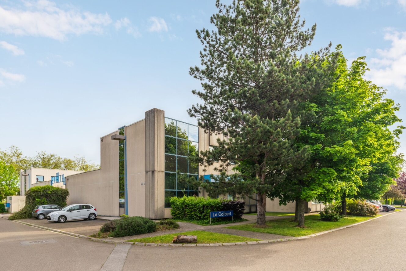 Immeuble de bureaux moderne avec de grandes fenêtres, entouré d'arbres et de verdure, avec des voitures garées et un panneau intitulé Le Colbert près de l'entrée.