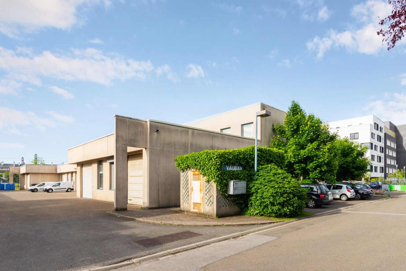 Bâtiment commercial de plain-pied à toit plat, entouré de voitures garées et de verdure, sous un ciel partiellement nuageux.