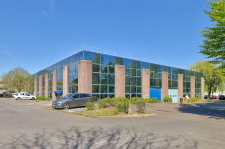 Un immeuble de bureaux moderne de deux étages avec de grandes fenêtres en verre réfléchissant, des colonnes en pierre, un parking environnant et plusieurs voitures garées sous un ciel dégagé.