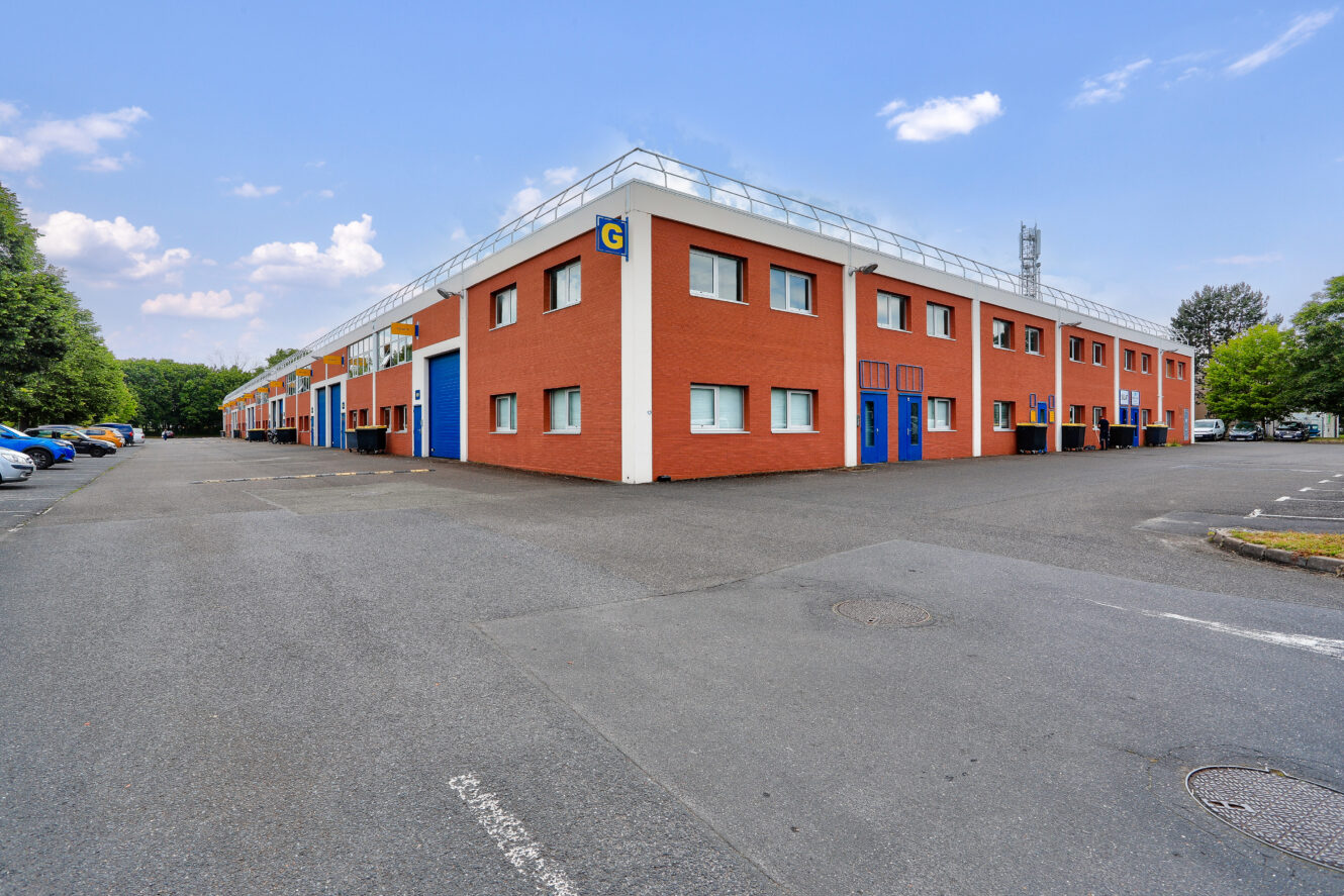 Un bâtiment commercial de deux étages en briques rouges avec des portes et des fenêtres bleues, portant la lettre G, situé sur un terrain pavé sous un ciel partiellement nuageux.